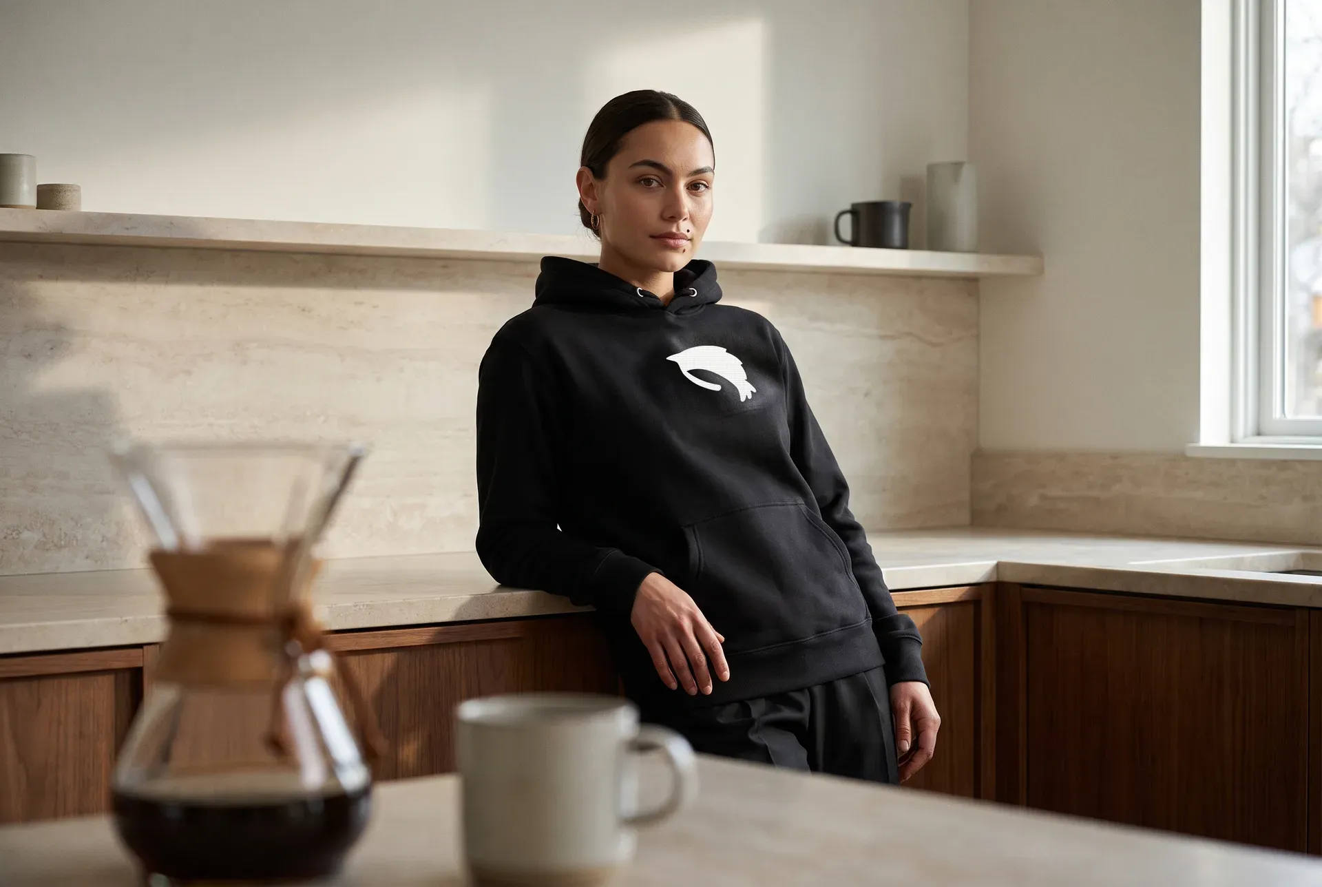 A woman in a dark hoodie with an embroidered shrimp motif, in a bright kitchen — quiet, worn shot.
