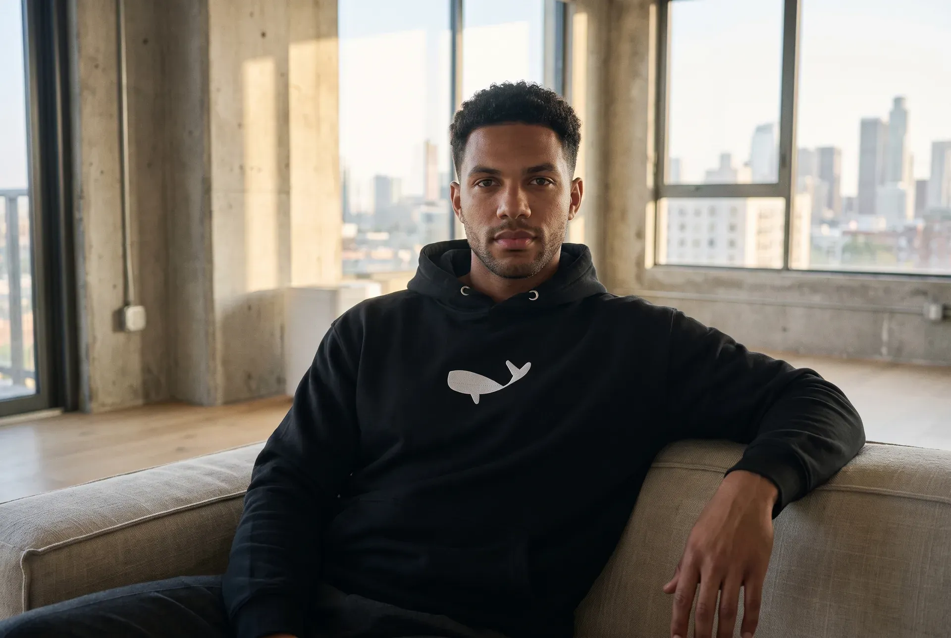 A man in a dark hoodie with an embroidered humpback whale on the chest, in a bright loft — quiet, worn shot.