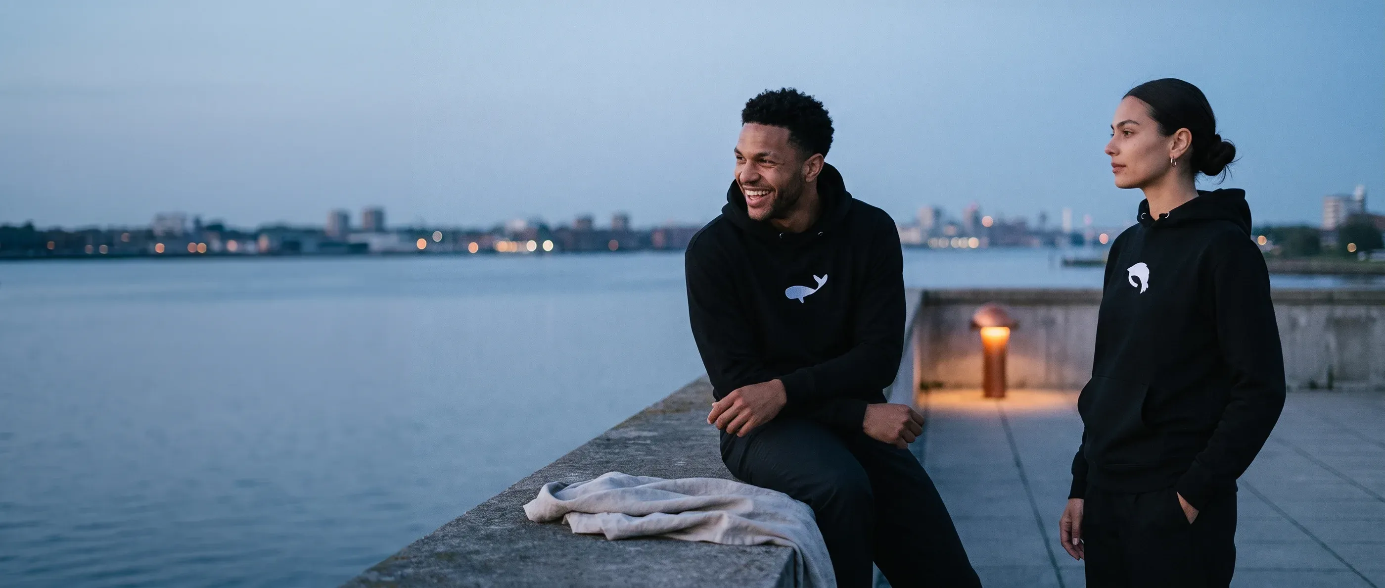 A person in a black whale hoodie at a floor-to-ceiling window at blue hour, calm water below