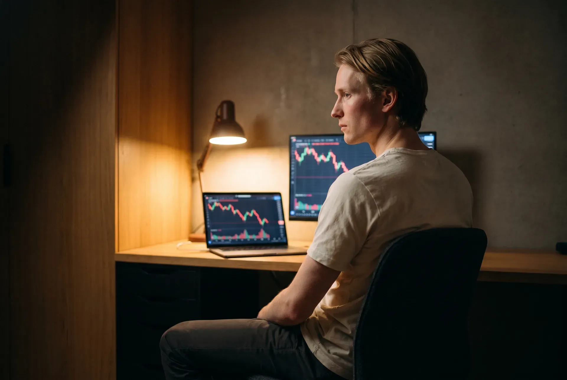 A young man in a light tee sits calmly at his desk before two screens showing falling red price charts, warm lamp light