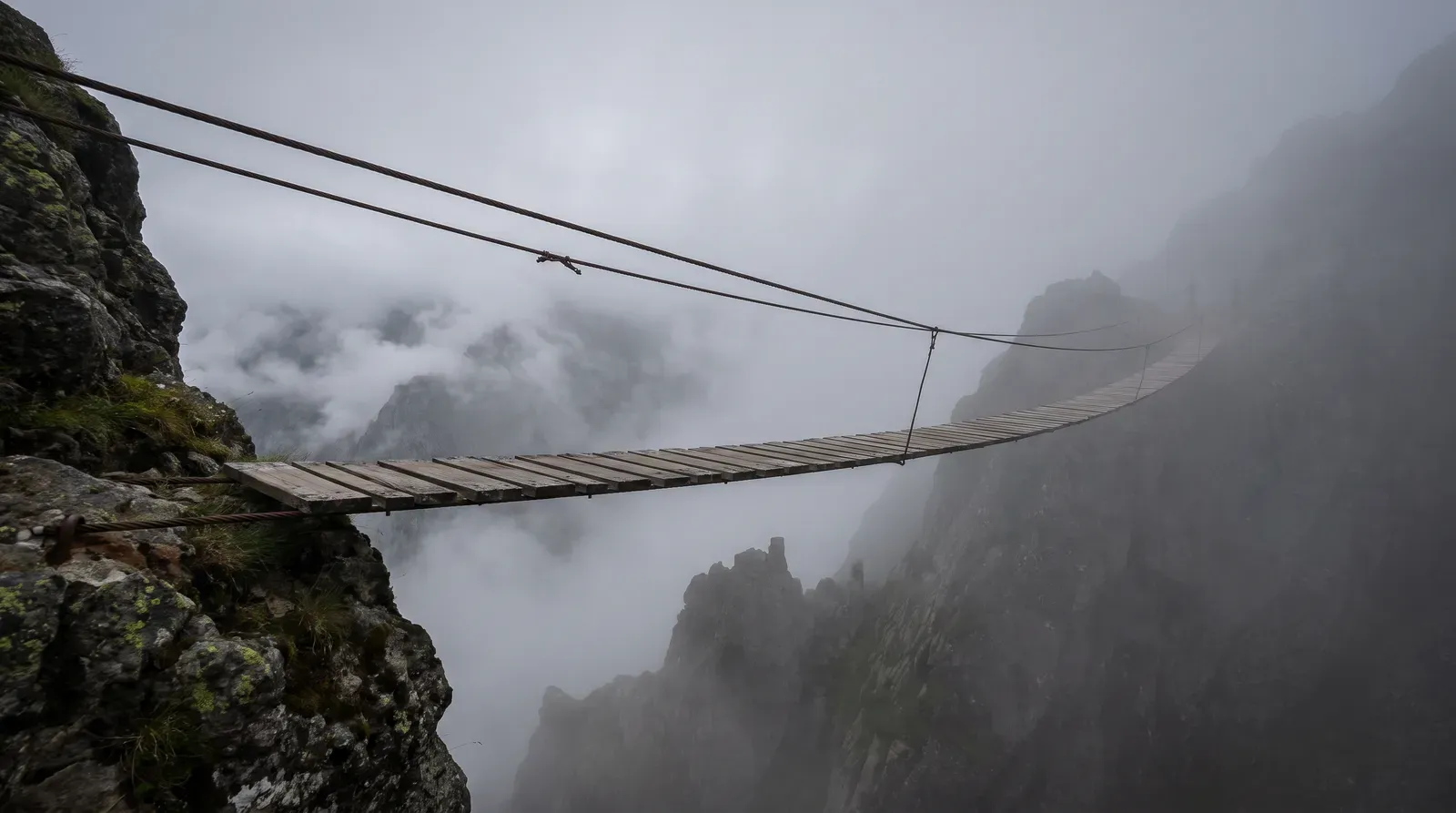 A narrow plank suspension bridge crossing a fog-filled gorge, the far end disappearing into grey