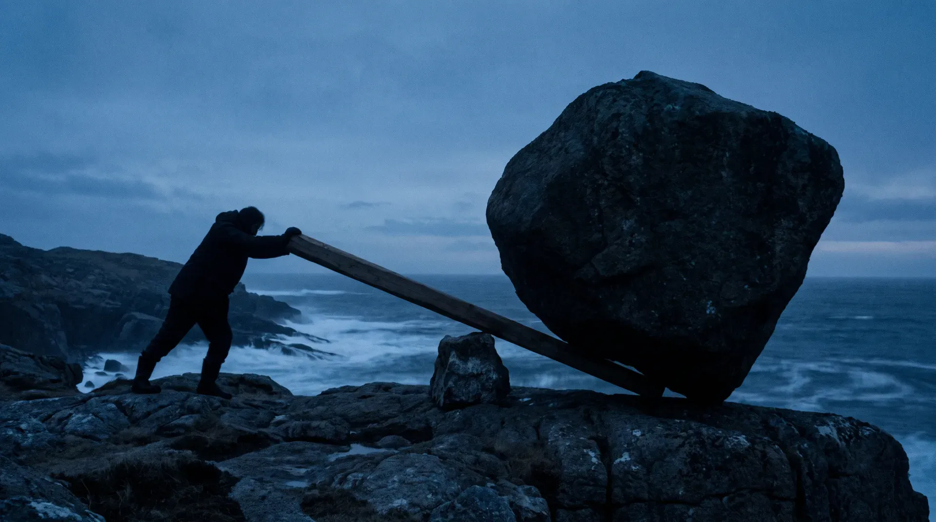 A person at dusk on a rocky coastline tipping an enormous boulder with a long wooden lever over a small fulcrum stone — great force, thin margin for error