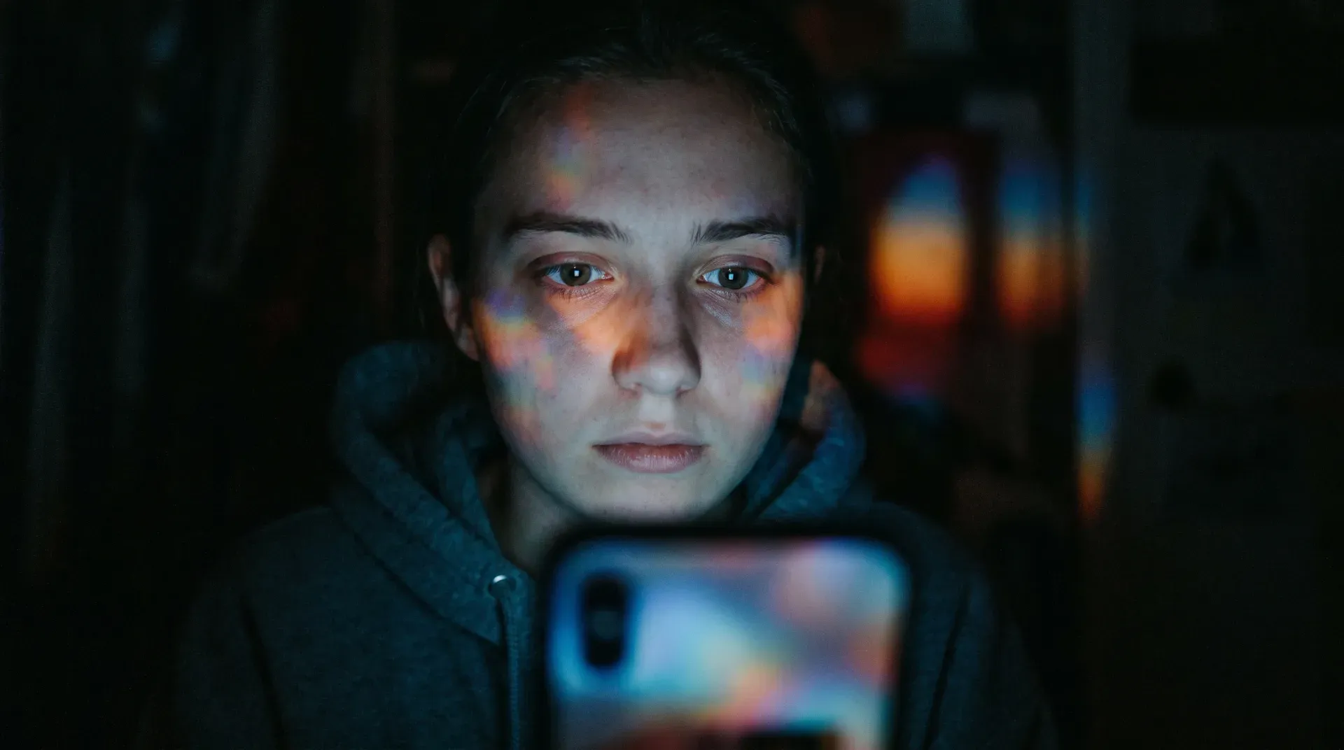 A young face lit only by the harsh glow of a smartphone, colorful reflections in the eyes, somewhere between excitement and exhaustion — three in the morning