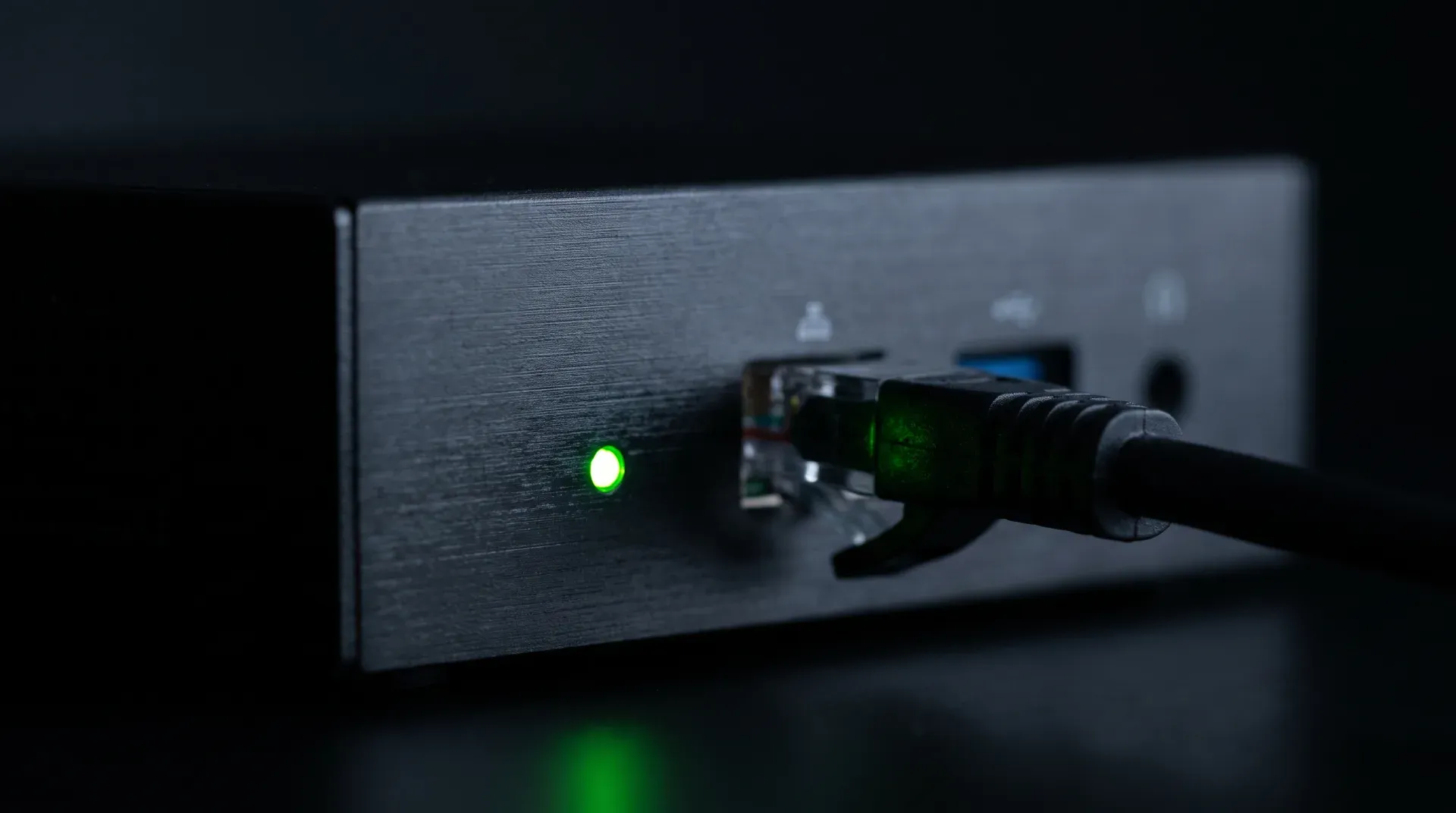 Macro shot of a small black mini computer in the dark: a single green status LED, next to it a connected ethernet cable